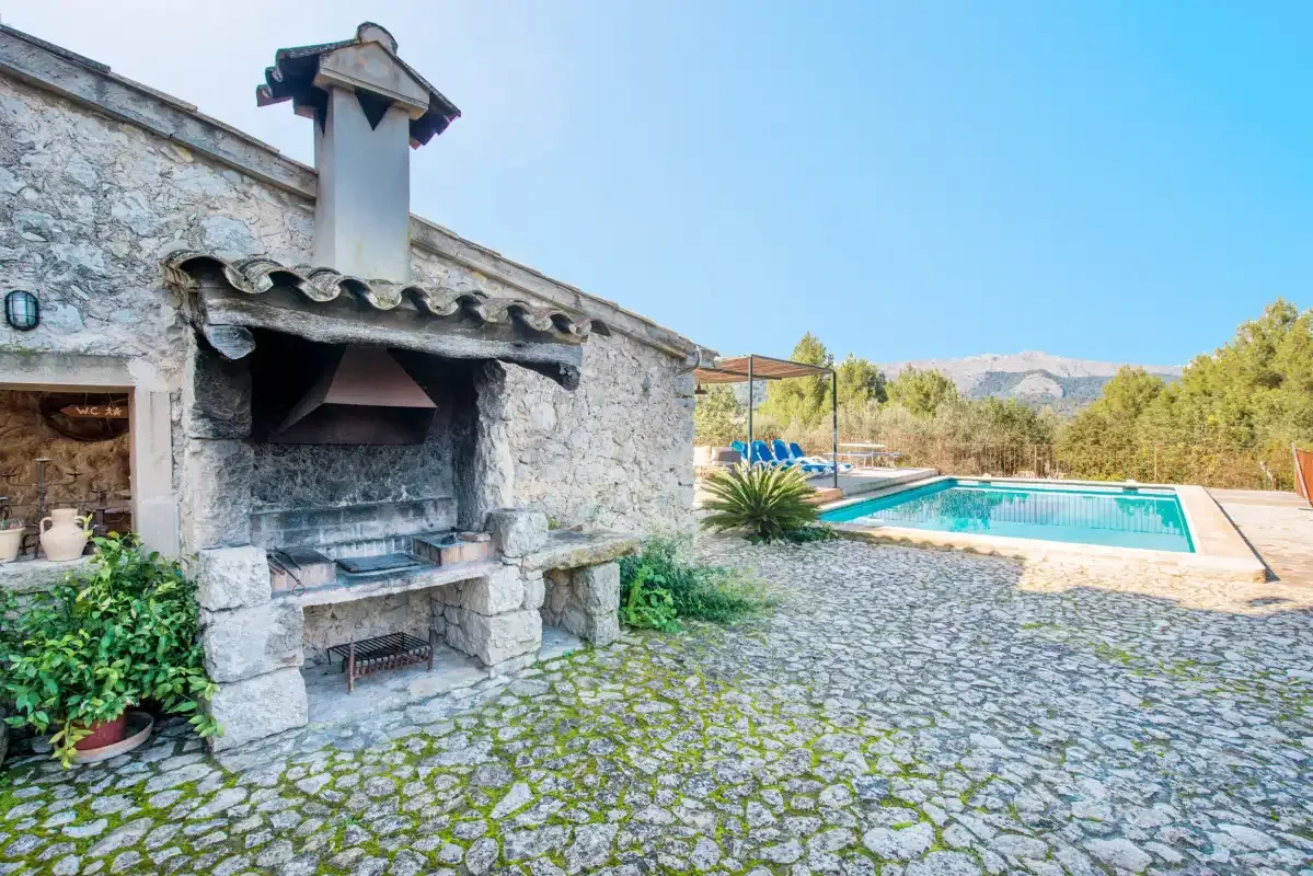 A stone outdoor barbecue stands beside a swimming pool in a paved courtyard.