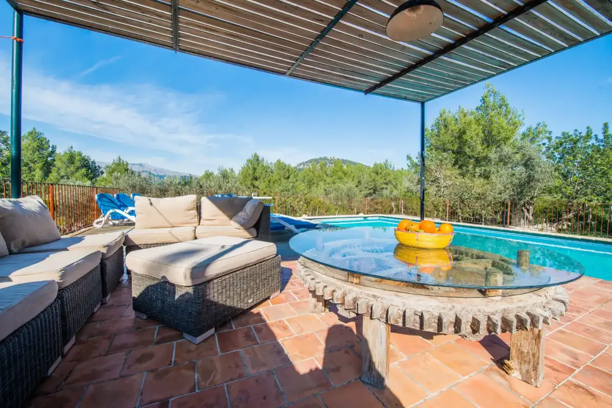 A covered outdoor patio has cushioned seating beside a round table and pool.