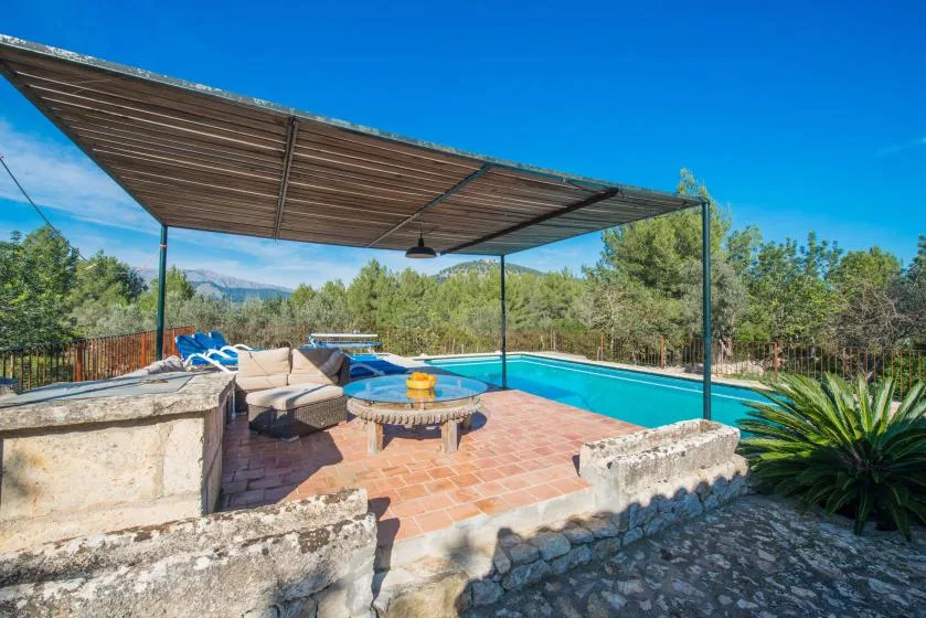 Un patio junto a la piscina tiene asientos de descanso bajo un amplio toldo.