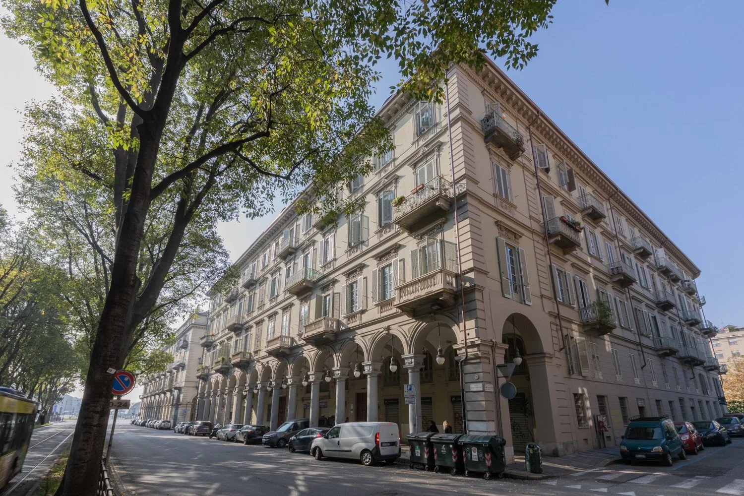 A city street lined with parked vehicles beside a multi-story building with arched ground-floor openings.