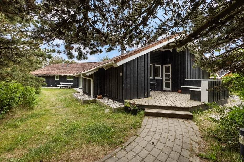 A black wooden house with a raised deck and a paved path through the yard.