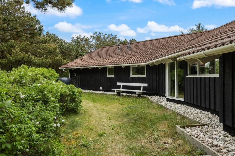 A dark wooden house stands beside a grassy yard and leafy shrubs.
