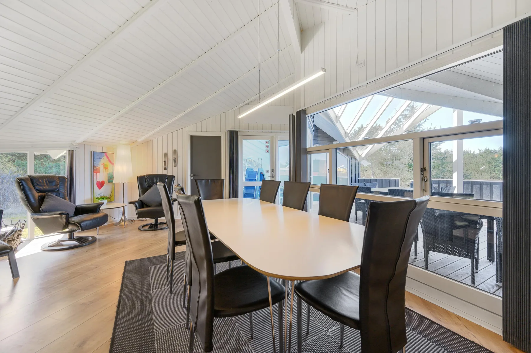 A dining area with a long table and dark seating beside large windows.