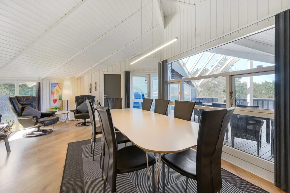 A bright dining area has a long table beside large windows overlooking a deck.