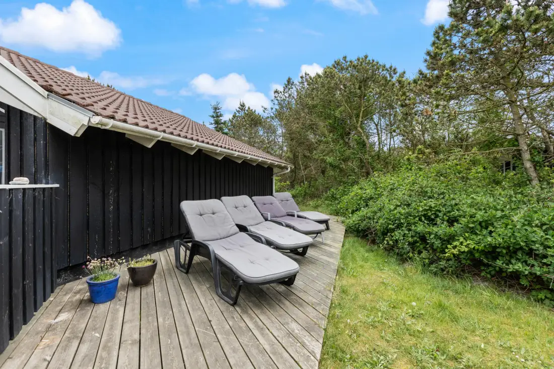An outdoor deck beside a dark wooden house has several lounge chairs and potted plants.