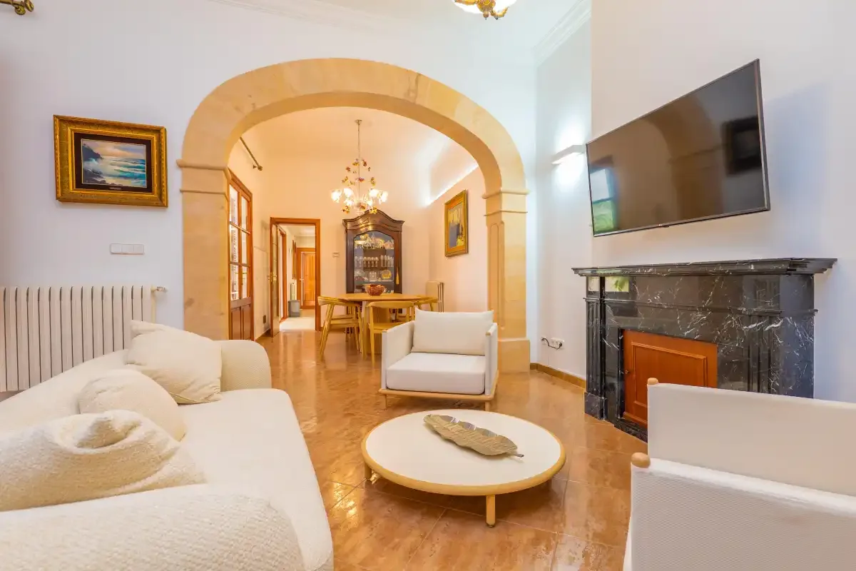 Una sala de estar con asientos claros, una chimenea y un comedor al fondo del arco.