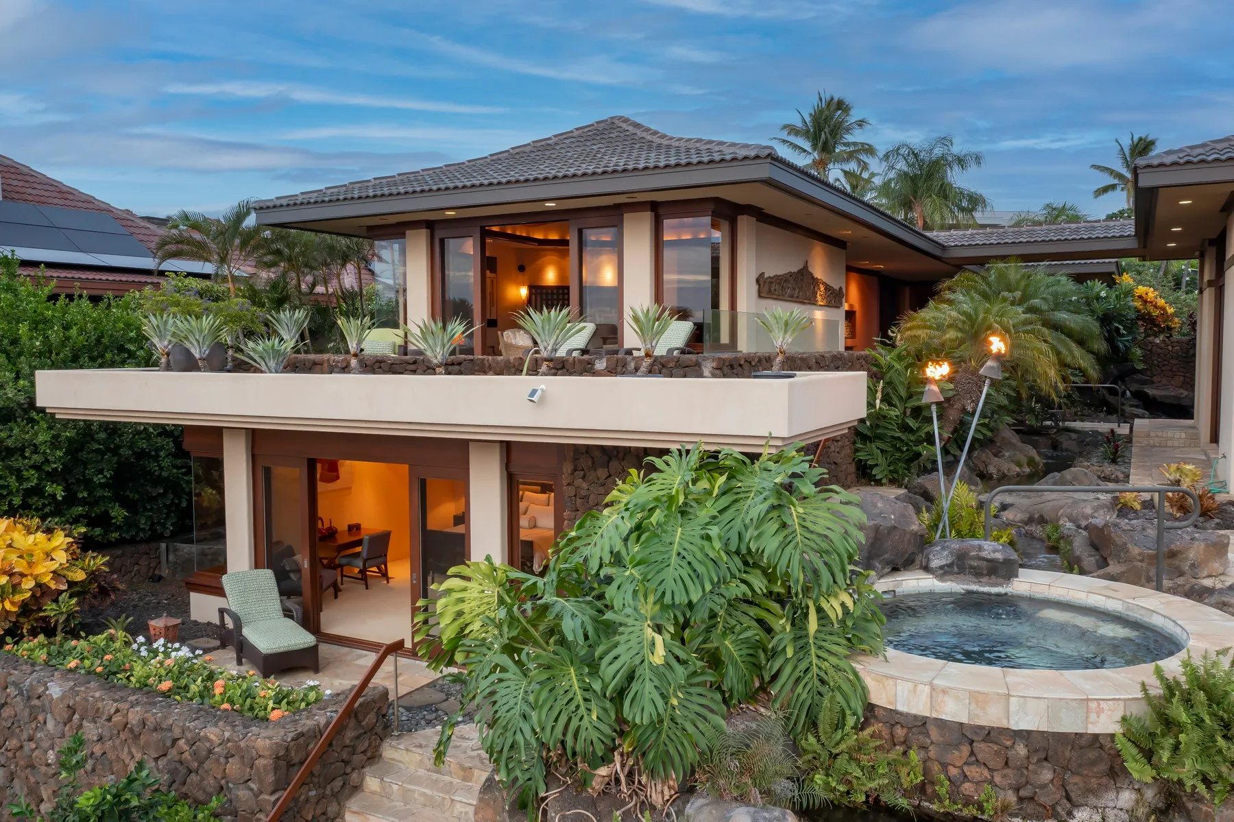 Casa tropical de dos plantas con terraza superior frente a un jardín ajardinado y una piscina redonda.