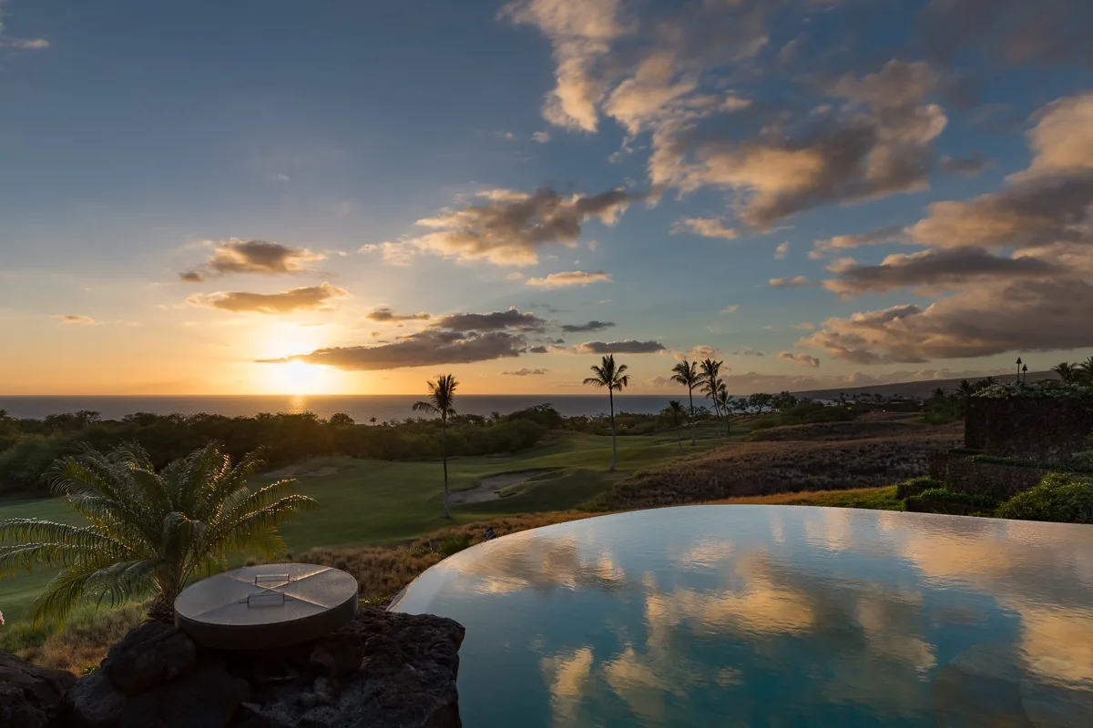Una piscina infinita refleja un atardecer colorido sobre un paisaje costero ajardinado.