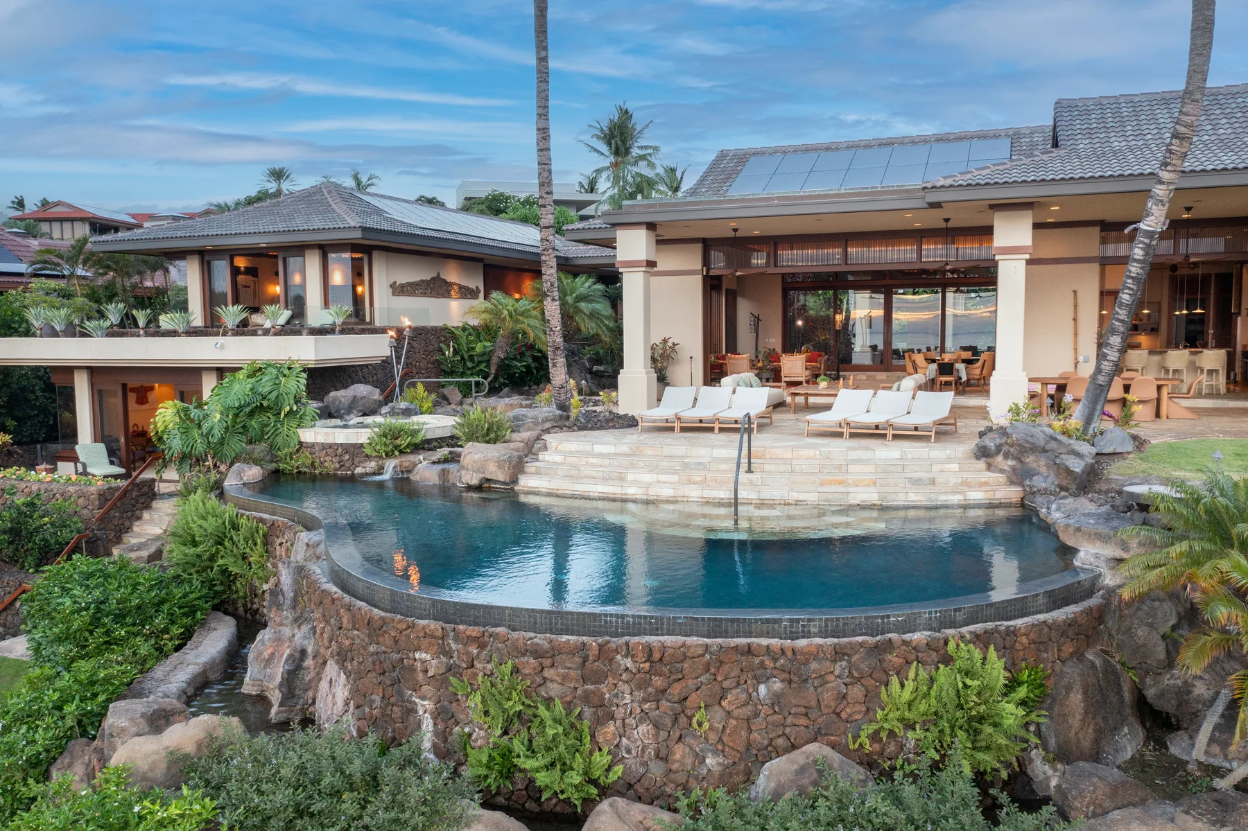 Una piscina al aire libre rodeada de jardines está frente a una casa con patio cubierto.