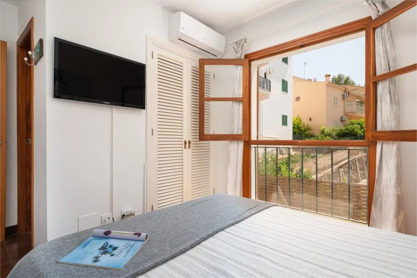 Un dormitorio con cama, televisor en la pared y ventanas abiertas con vistas a edificios cercanos.