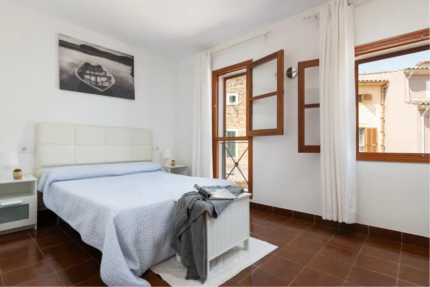 Un dormitorio luminoso con una cama doble, mesitas de noche y ventanas con vistas a edificios cercanos.