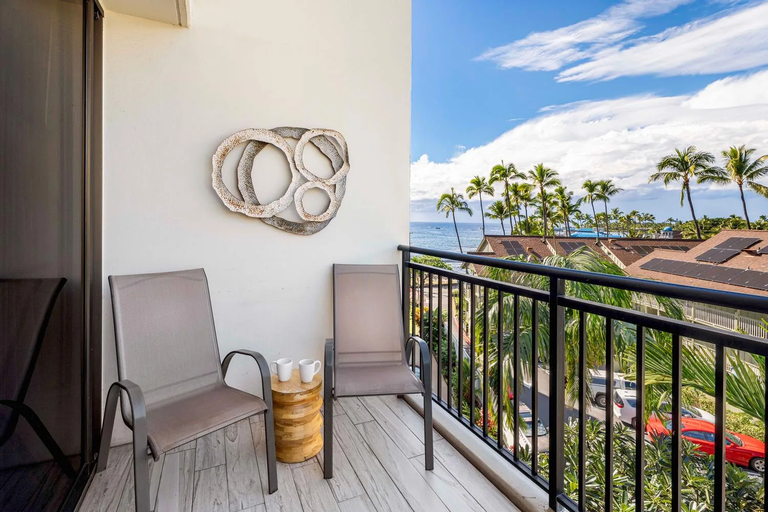 A balcony with two chairs overlooks palm trees and nearby rooftops.