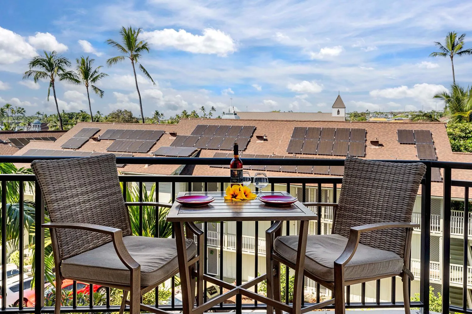 A balcony dining area with a table and two woven seats overlooks palm trees and rooftops.