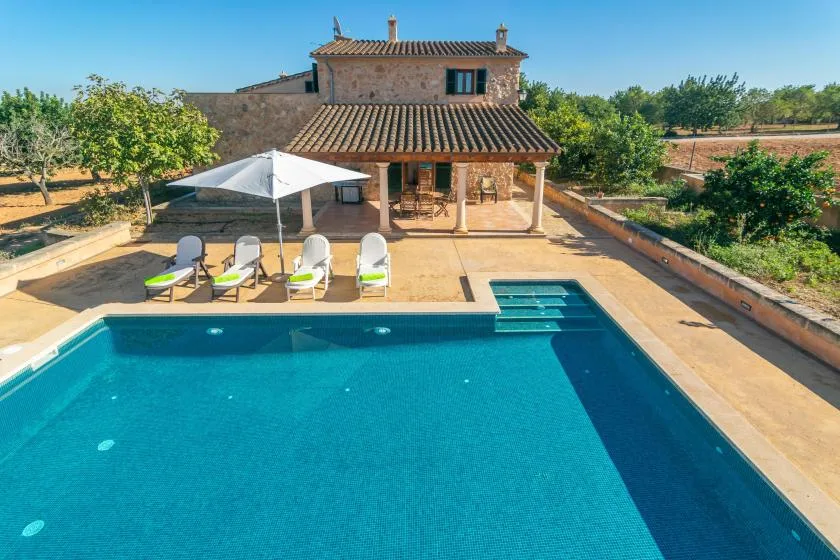 Una piscina y un patio están frente a una casa de piedra en un jardín cuidado.