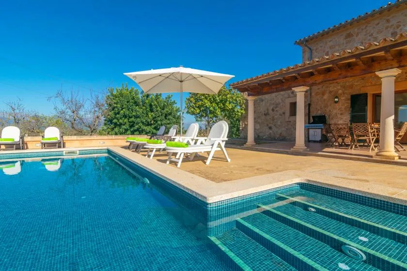 Una piscina al aire libre junto a una casa de piedra, con asientos bajo una sombrilla grande.