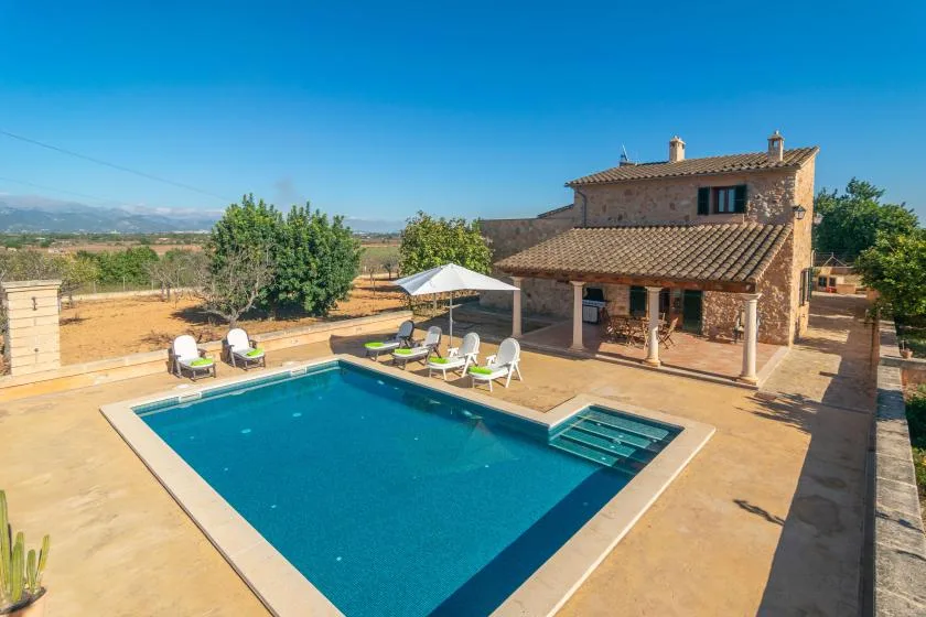 Una piscina y un patio están junto a una casa de piedra en una escena soleada al aire libre.