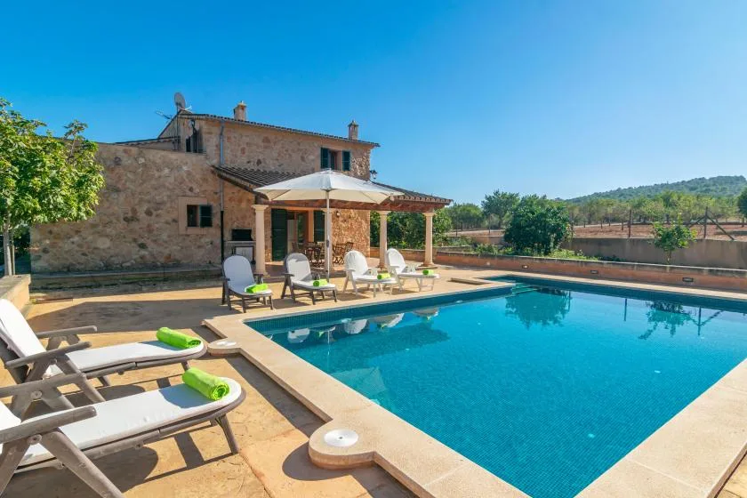 Piscina al aire libre junto a una casa de piedra, con una zona de estar sombreada.