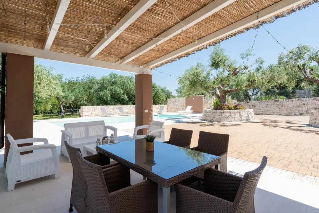 Una zona de comedor exterior a la sombra da a una piscina y un jardín ajardinado.