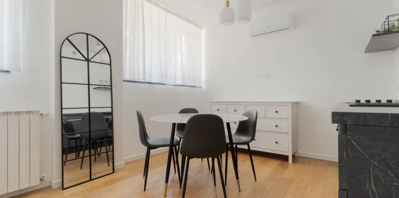 Un comedor luminoso con mesa, asientos y un espejo alto con arco.