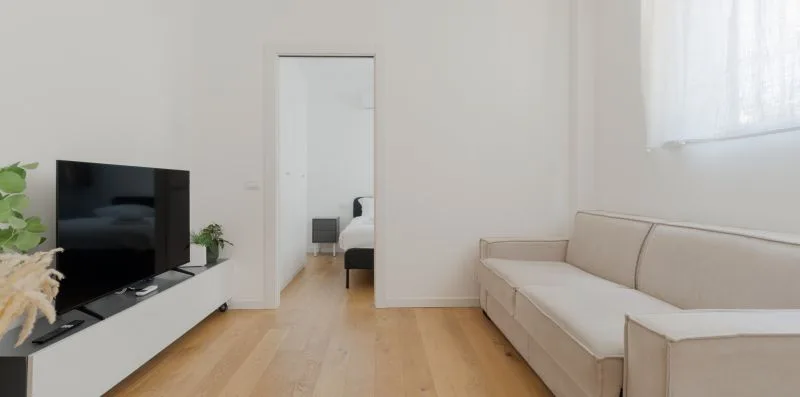 Una sala luminosa con televisor, asientos claros y una puerta hacia un dormitorio.