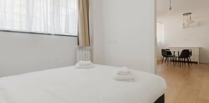 Un dormitorio luminoso con una cama bien arreglada y un comedor al fondo.