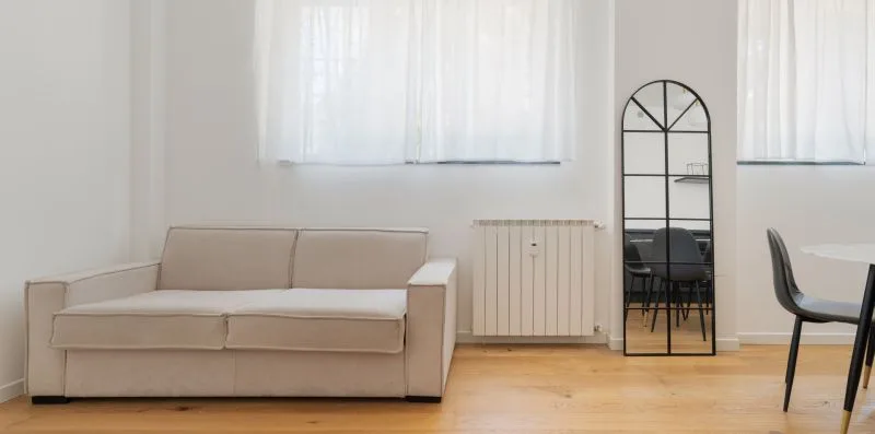 Una sala luminosa con un sofá beige, un radiador y una mesa de comedor.