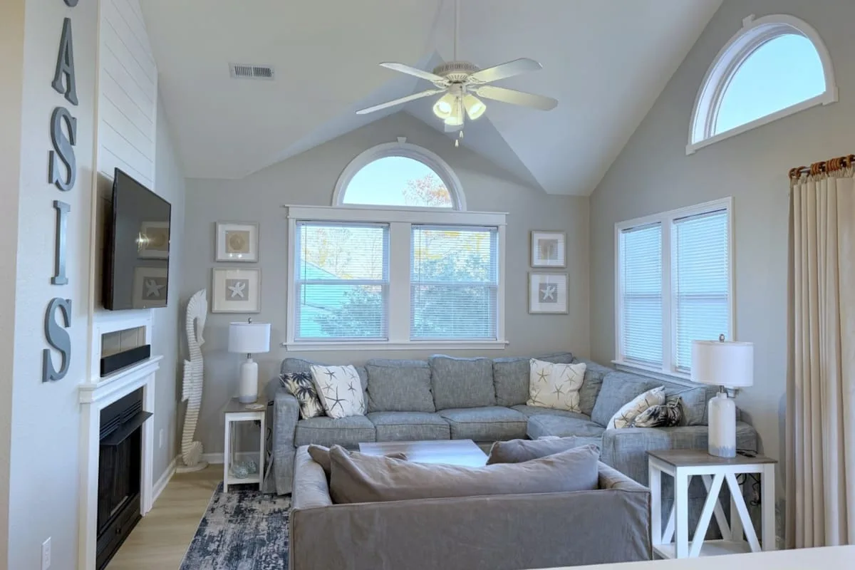 A bright living room with gray seating, large windows, and a ceiling fan.