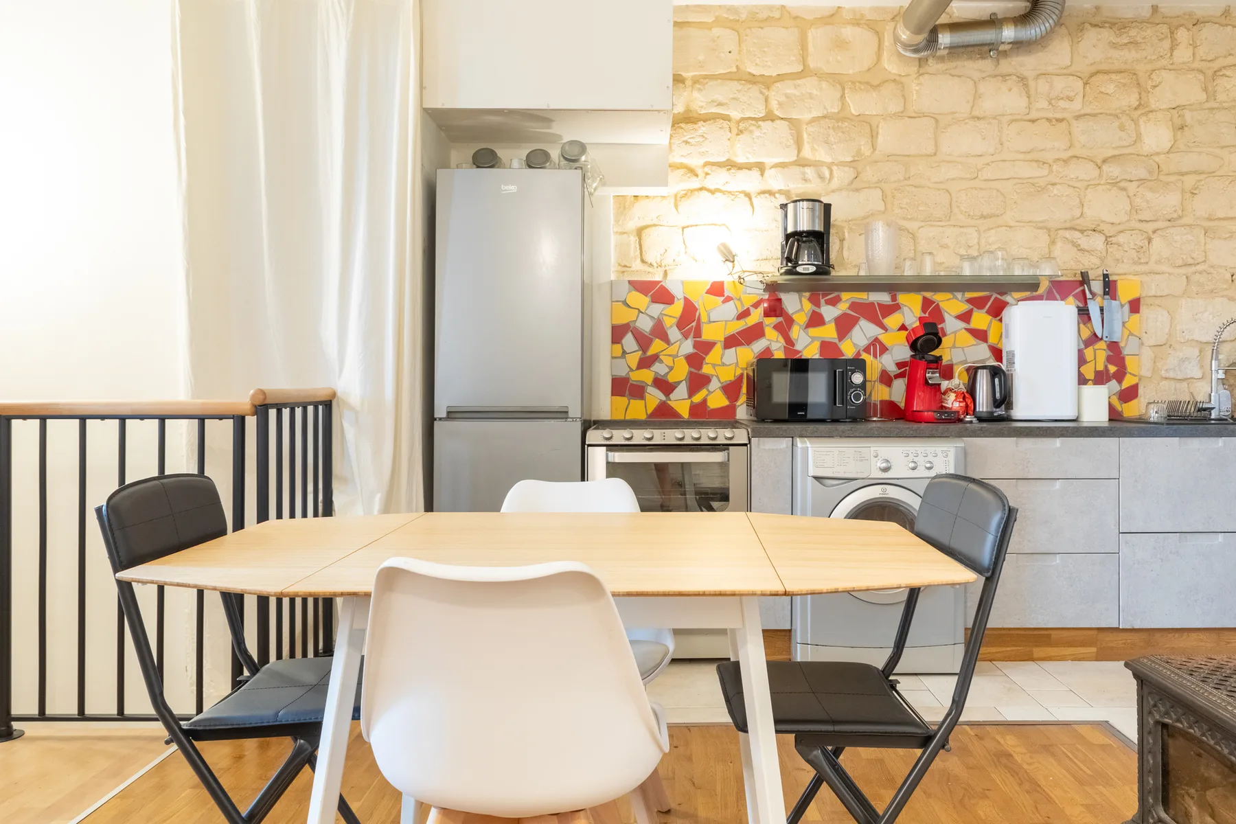 A dining table and chairs sit in a kitchen with colorful tile behind the counter.