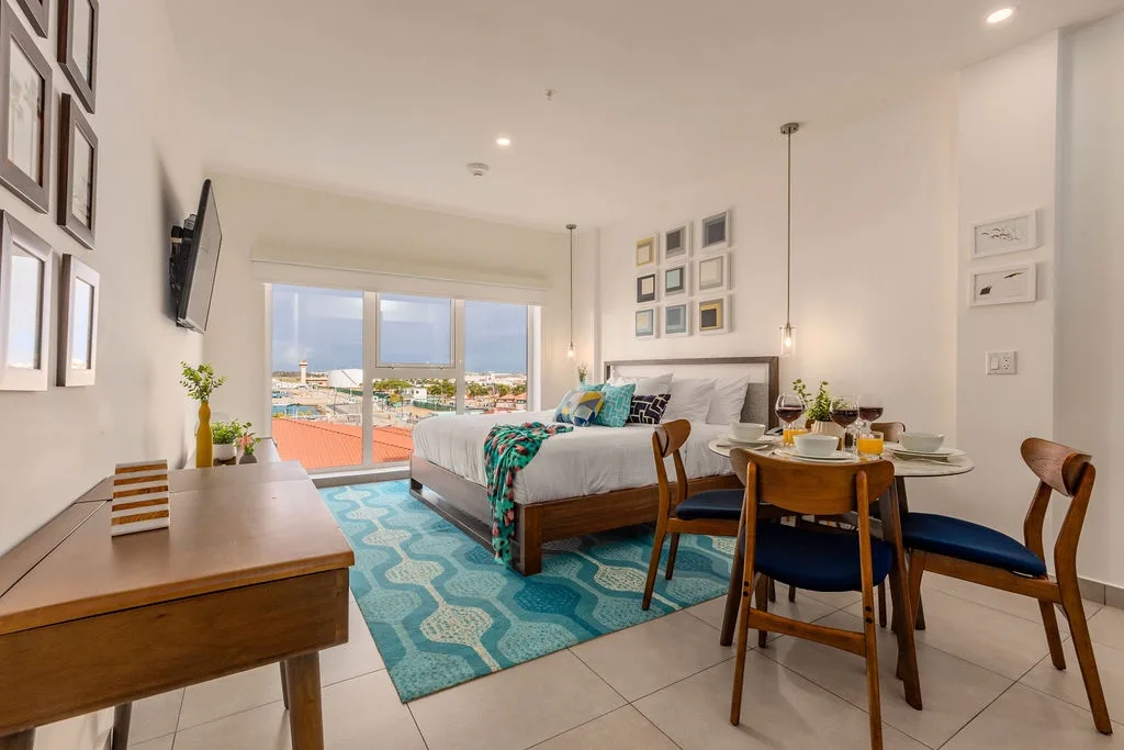 Your bright, open studio combines cozy sleeping space with a dining area for four, all bathed in natural light from floor-to-ceiling windows overlooking colorful rooftops and distant cityscape views.