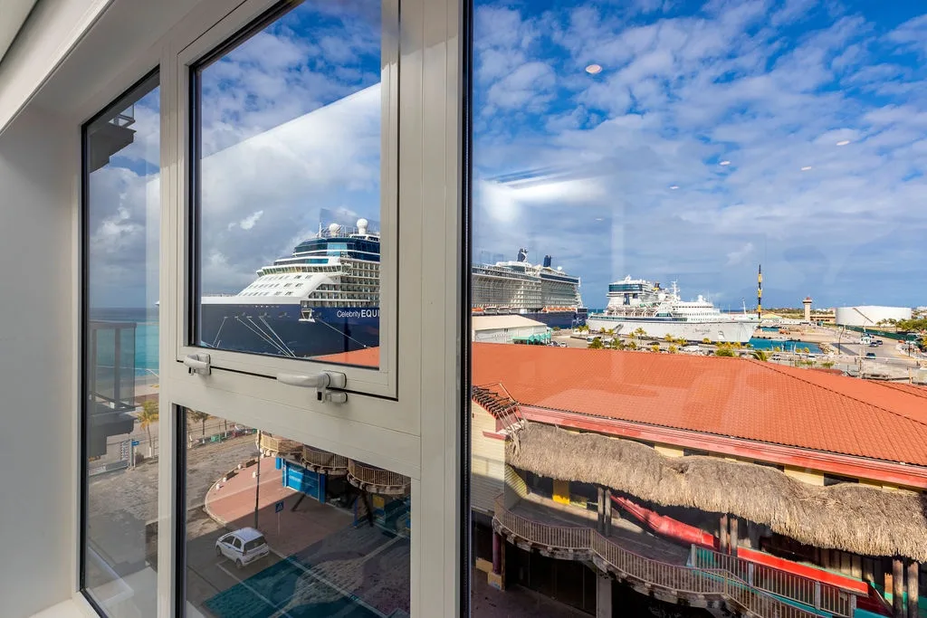 A view through the window reveals large cruise ships docked at the port, with colorful rooftops and coastal scenery visible in the distance.