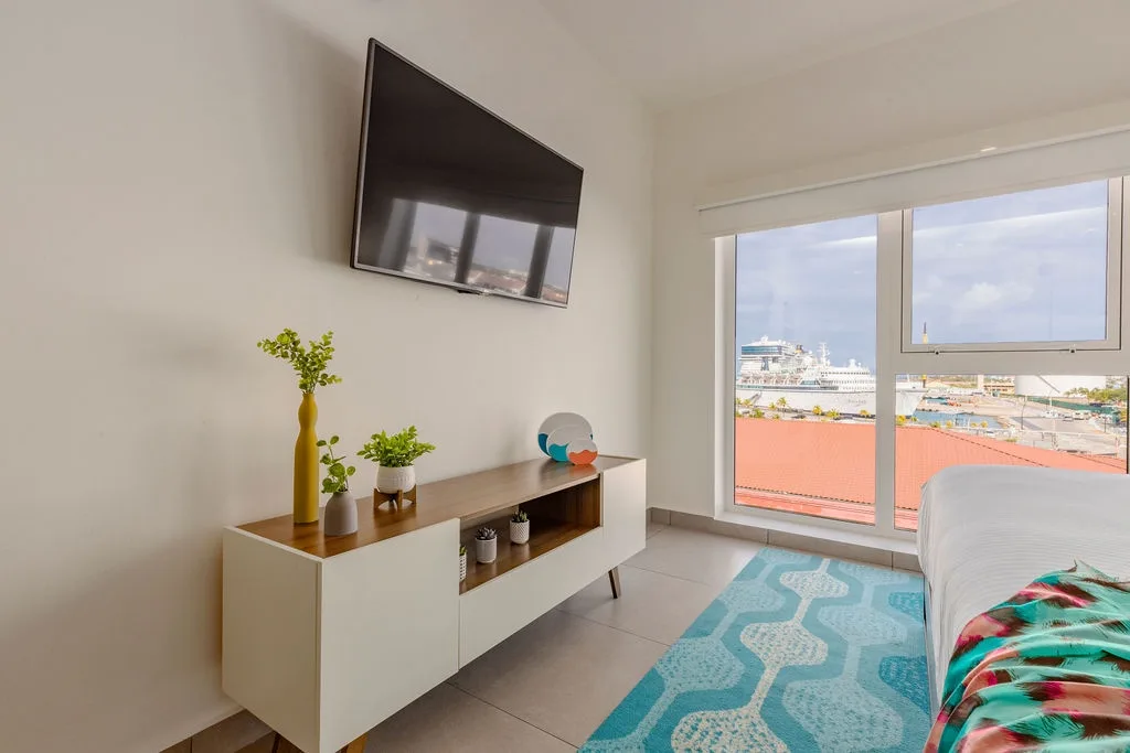 Your room features a wall-mounted TV and modern console, with bright windows framing a colorful cityscape and cruise ship views below.