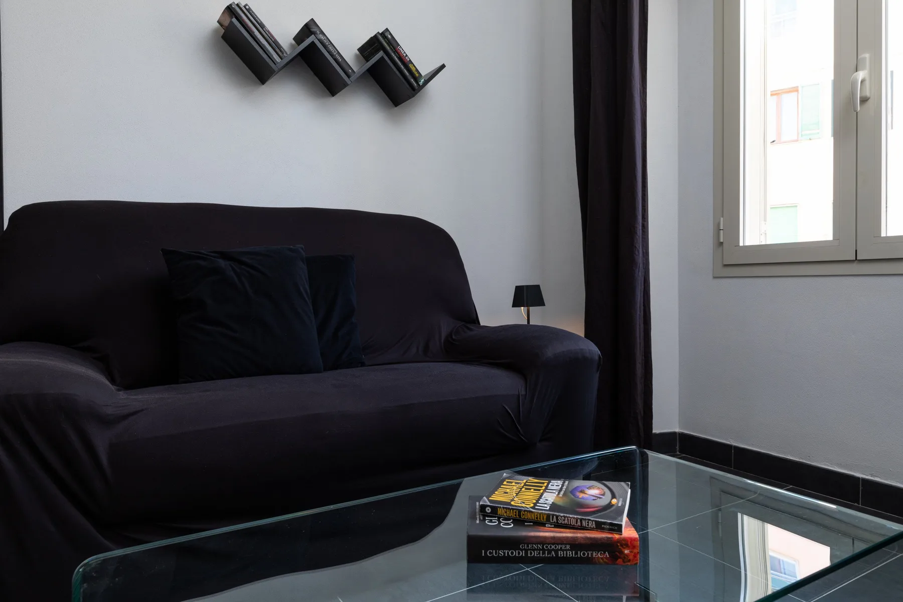 Un sofá oscuro está junto a una ventana, con libros sobre una mesa de centro de vidrio.