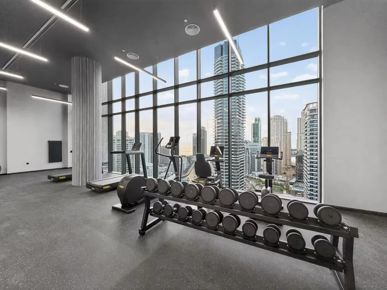 A fitness room with exercise equipment and floor-to-ceiling windows overlooking city buildings.