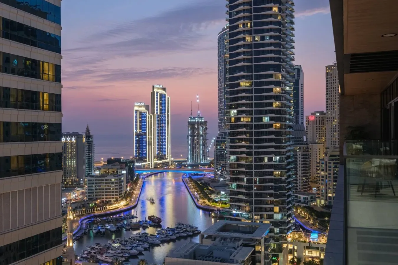 A city skyline at dusk overlooks a canal lined with illuminated buildings.