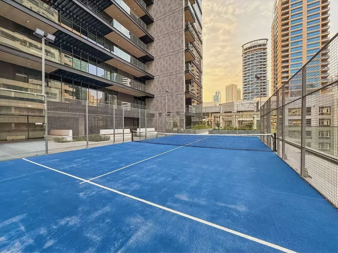 A blue outdoor court is enclosed by fencing between high-rise buildings.