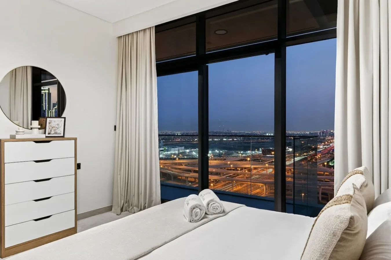 A bedroom with a bed beside a large window overlooking an illuminated city at dusk.
