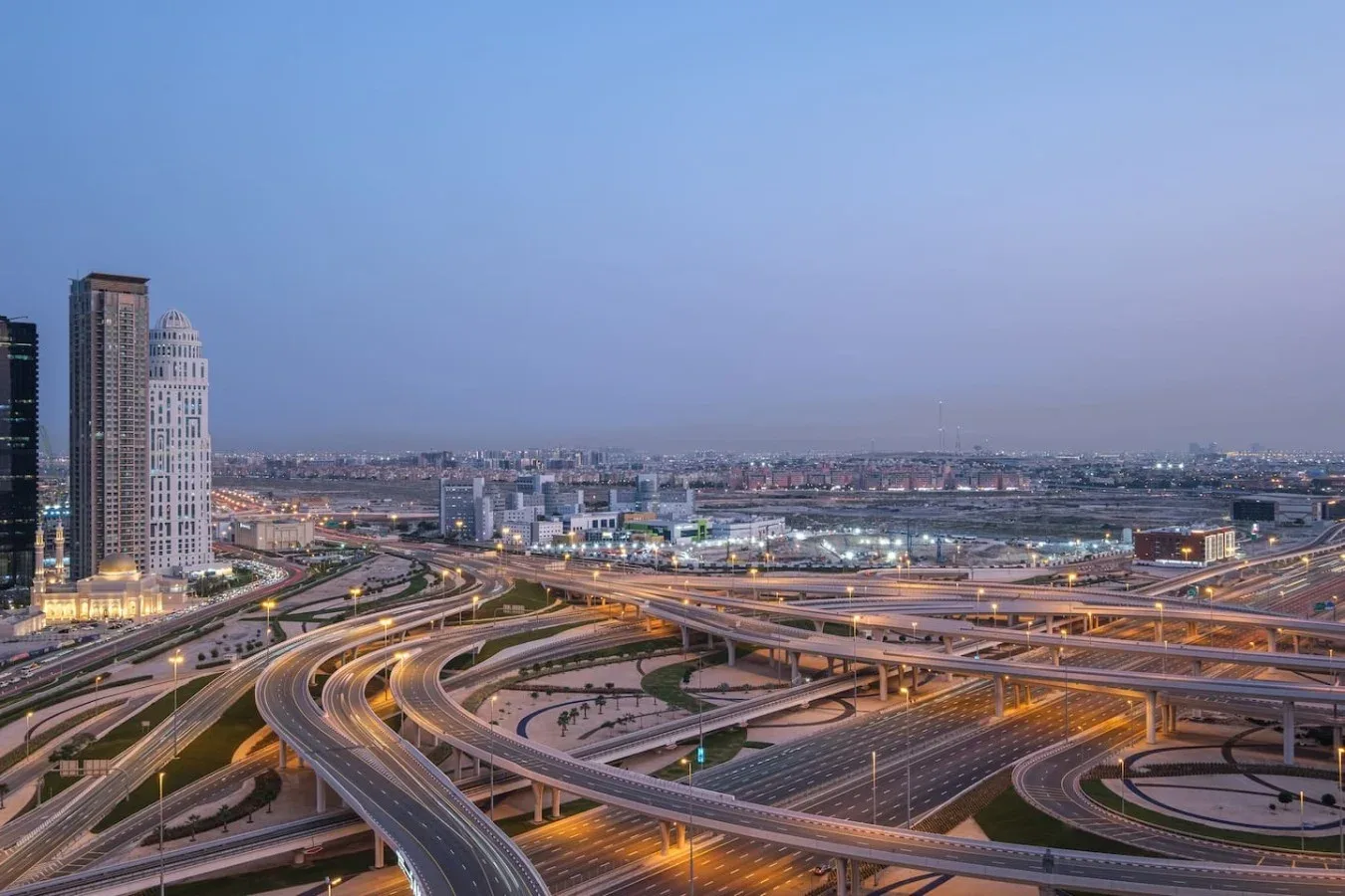 A city skyline rises behind a network of illuminated highway interchanges at dusk.