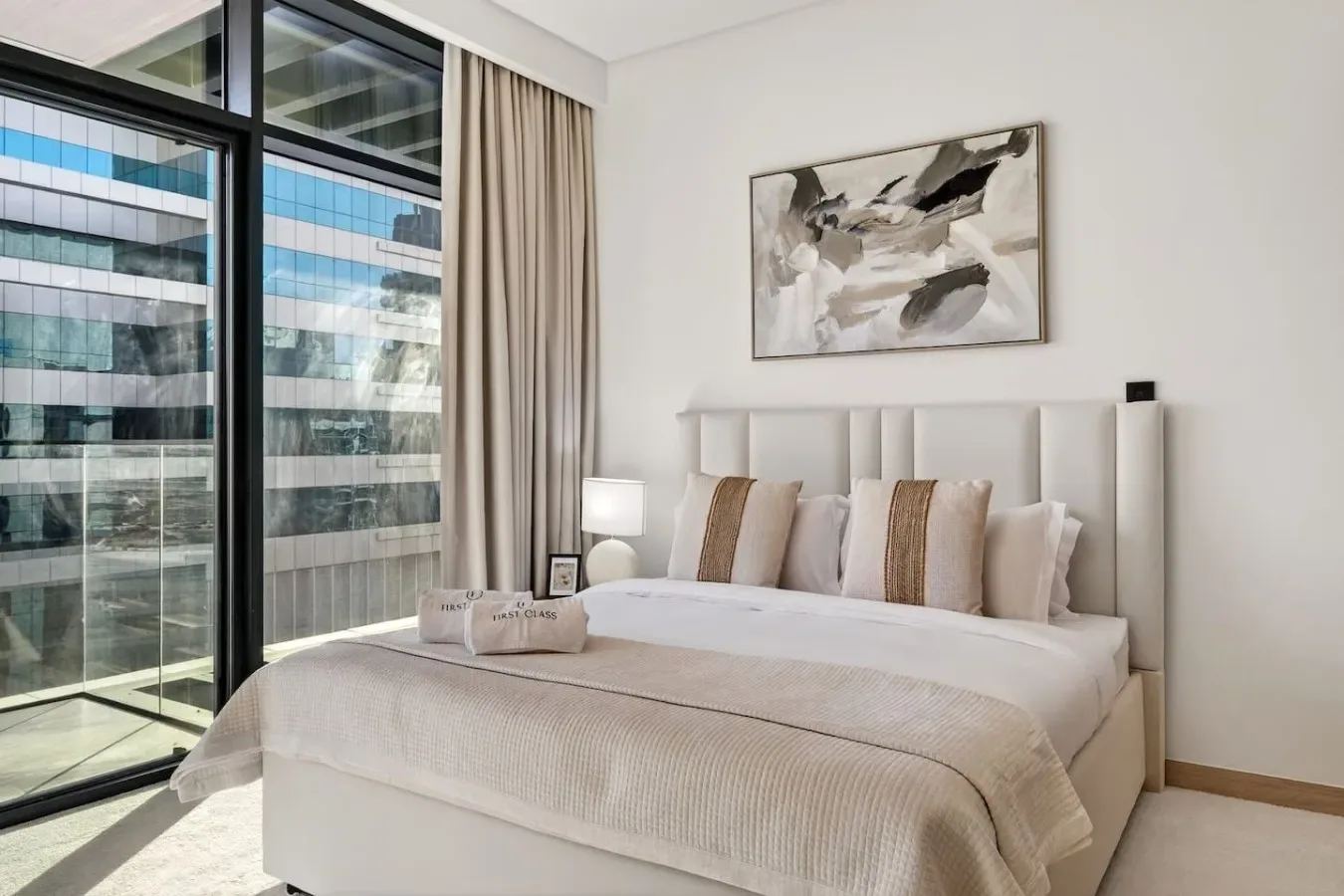 A bedroom with a neatly made bed, neutral bedding, and a large window.