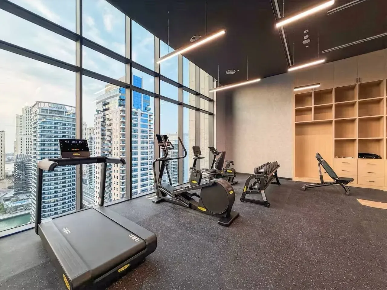 A gym with exercise equipment beside floor-to-ceiling windows overlooking nearby buildings.