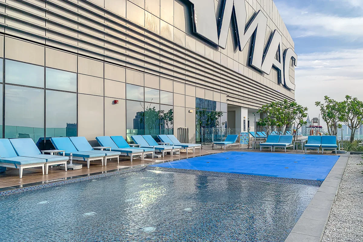 An outdoor swimming pool beside a modern building, with blue lounge seating along the edge.