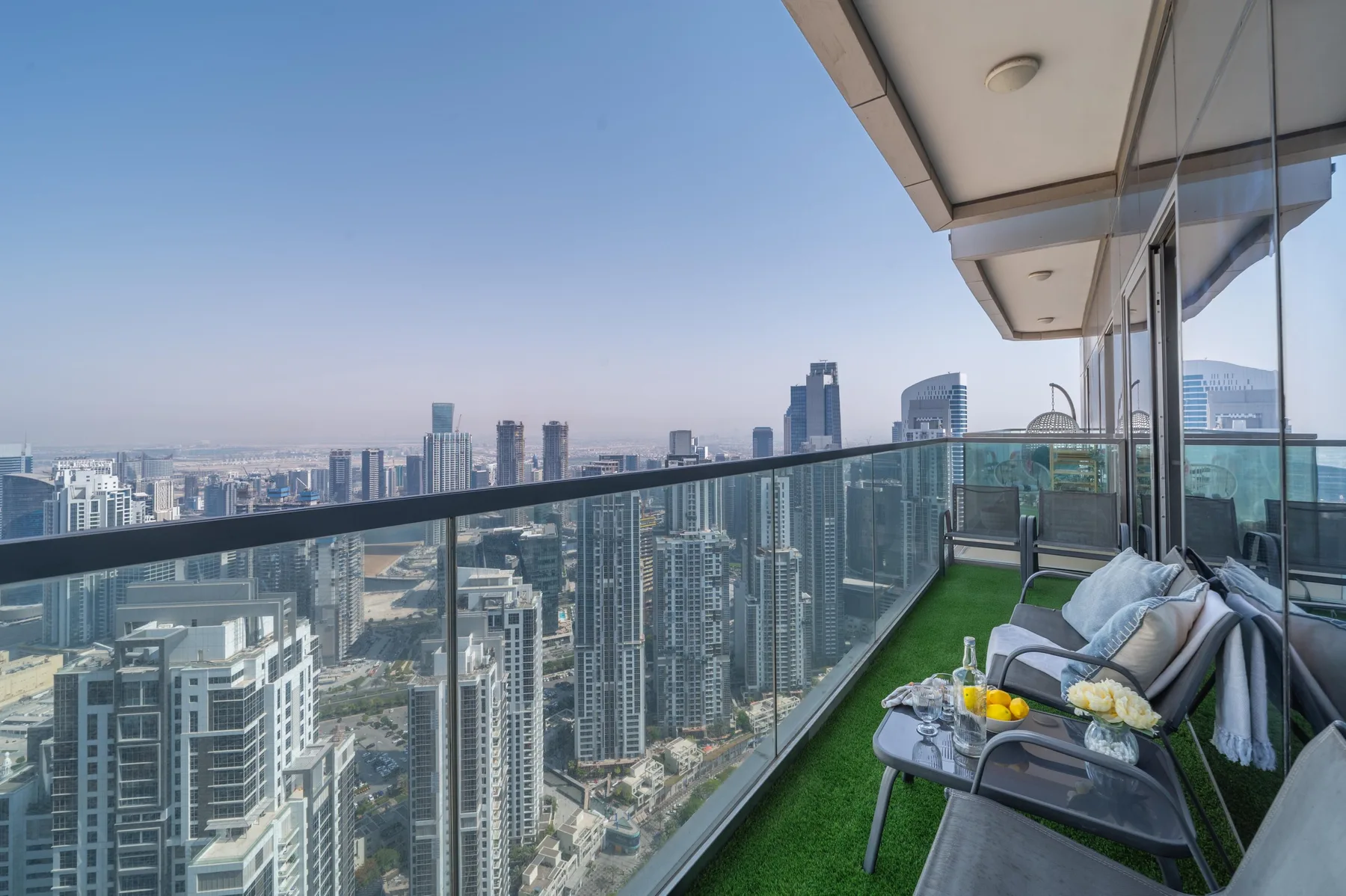 A high-rise balcony with glass railings, outdoor seating, and a city skyline view.