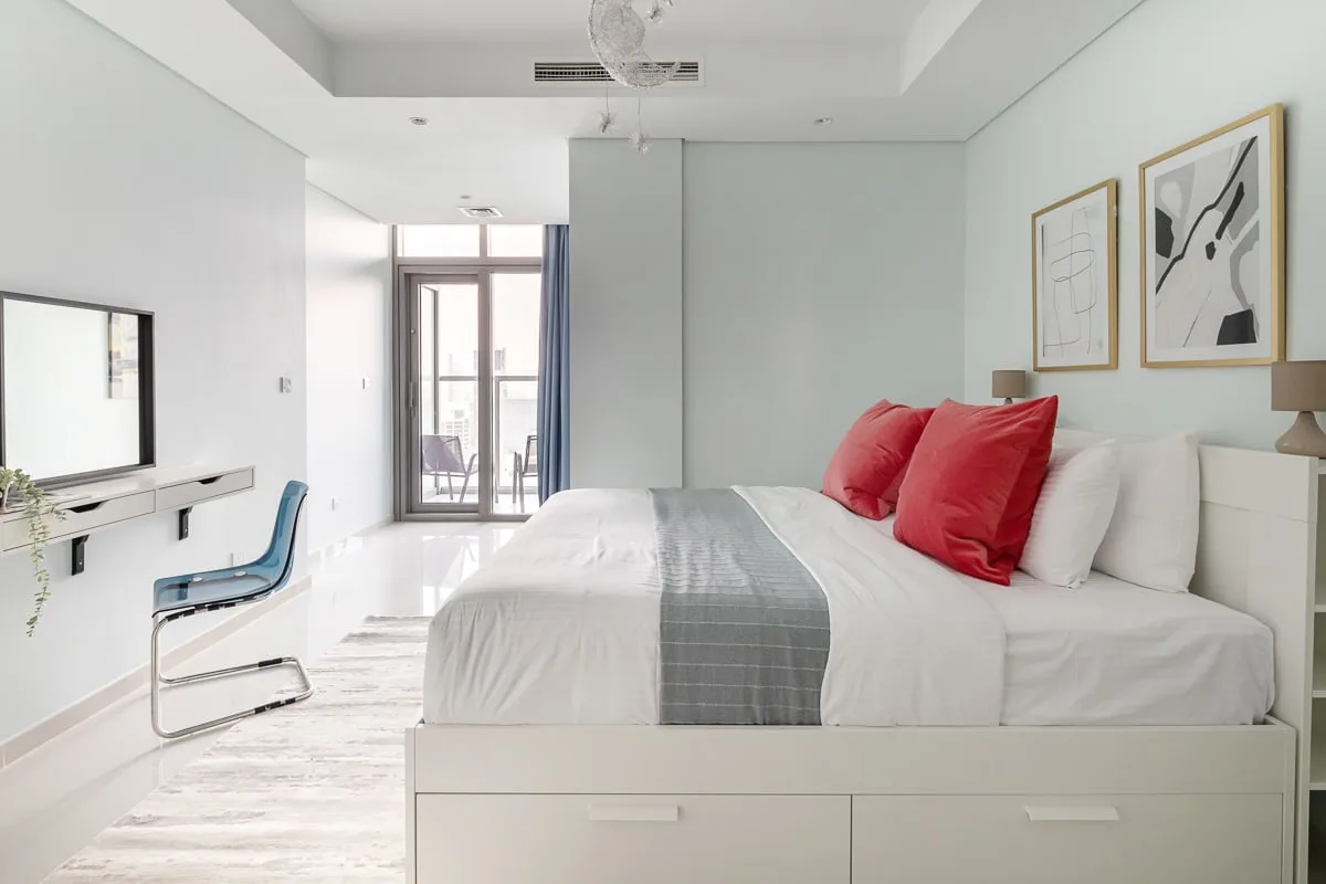 A bright bedroom features a bed with red pillows and a small desk.