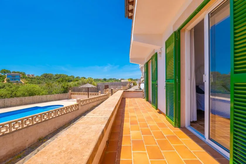 Una terraza soleada bordea un edificio con contraventanas verdes y da a una piscina.