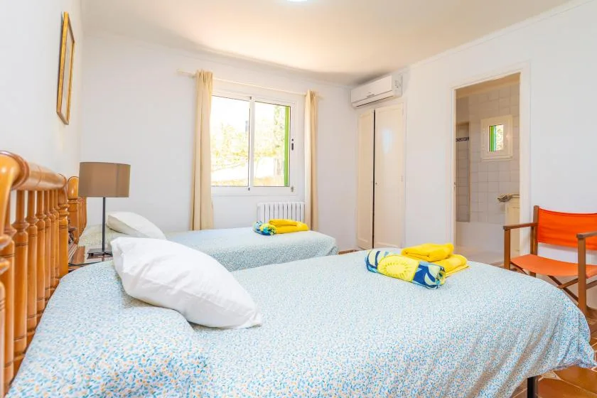Un dormitorio luminoso con dos camas, una ventana y una puerta a un baño con azulejos.