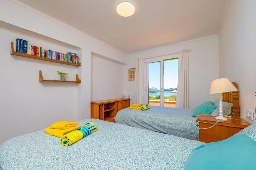 Un dormitorio luminoso con dos camas, estantes en la pared y una ventana con vista al agua.