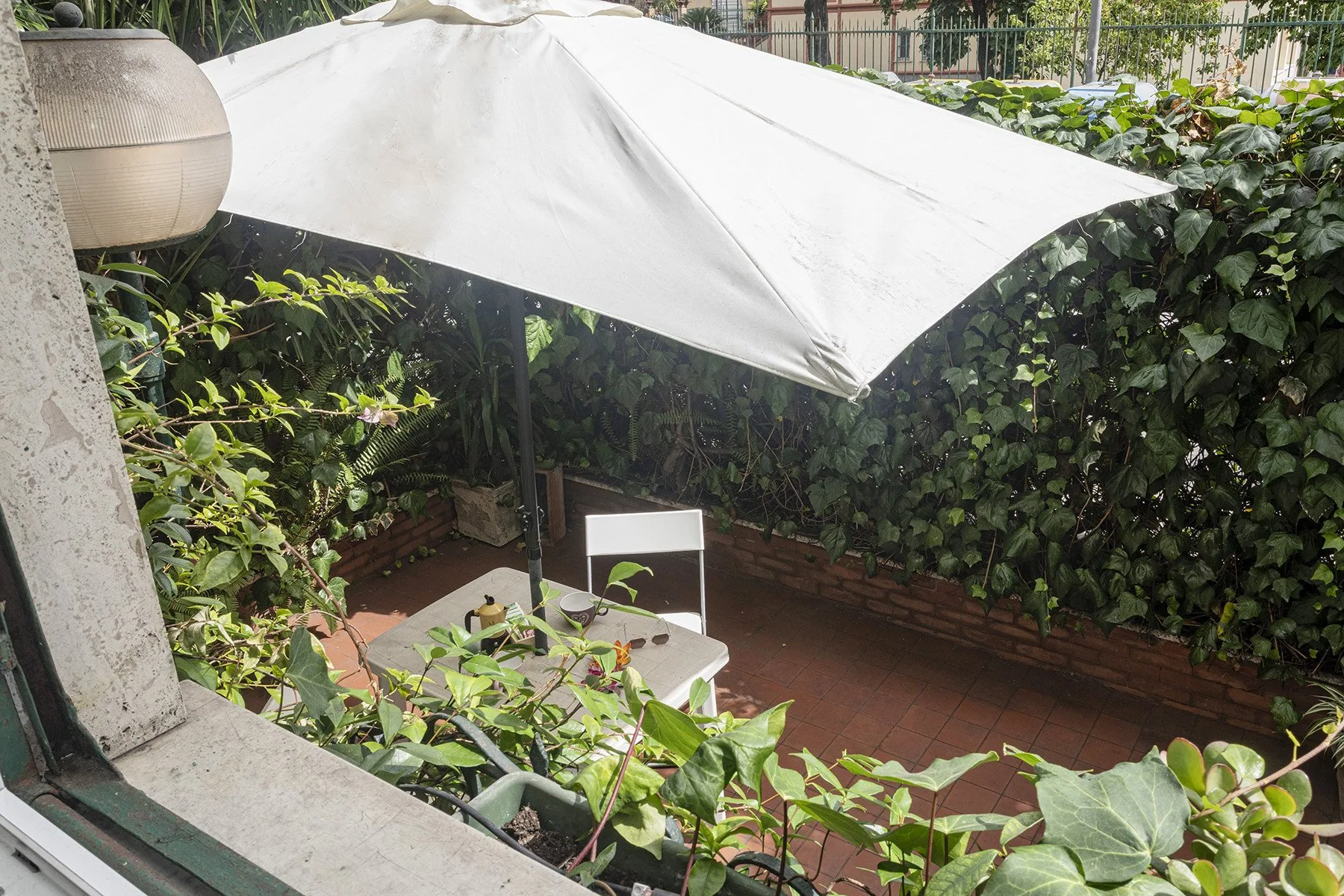 Un pequeño patio con asientos bajo una gran sombrilla blanca, rodeado de vegetación.