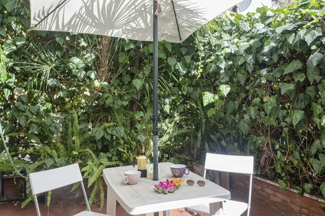 Un patio al aire libre con una mesa y sillas bajo una sombrilla grande, rodeado de vegetación.