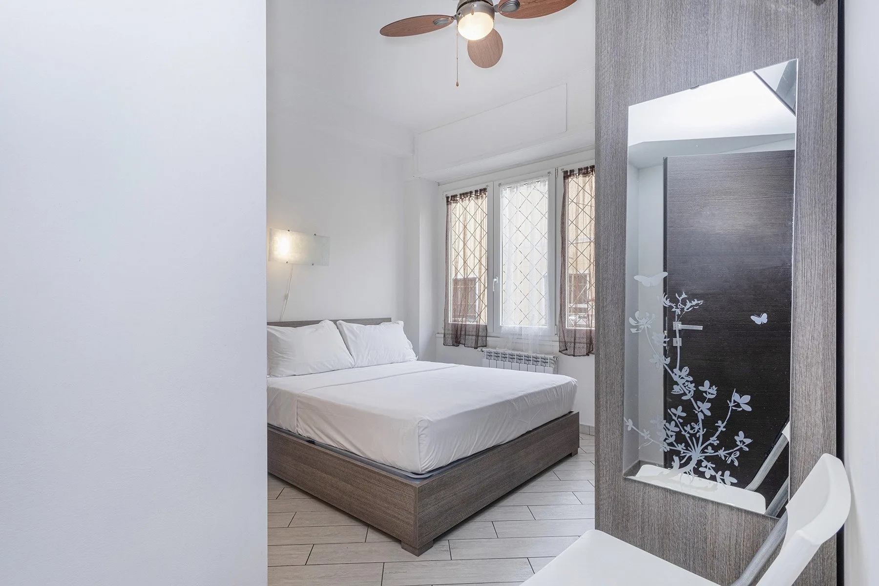 Un dormitorio luminoso con una cama, ventanas altas y un espejo de pared.