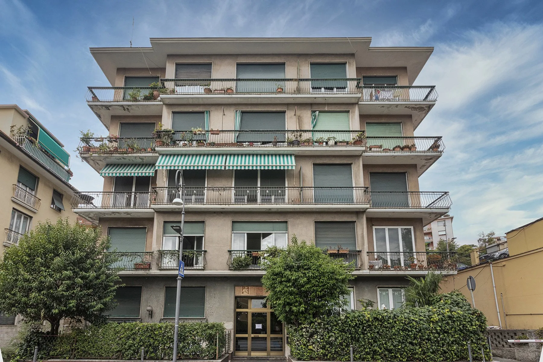 Un edificio residencial de varias plantas con balcones y vegetación junto a la calle.