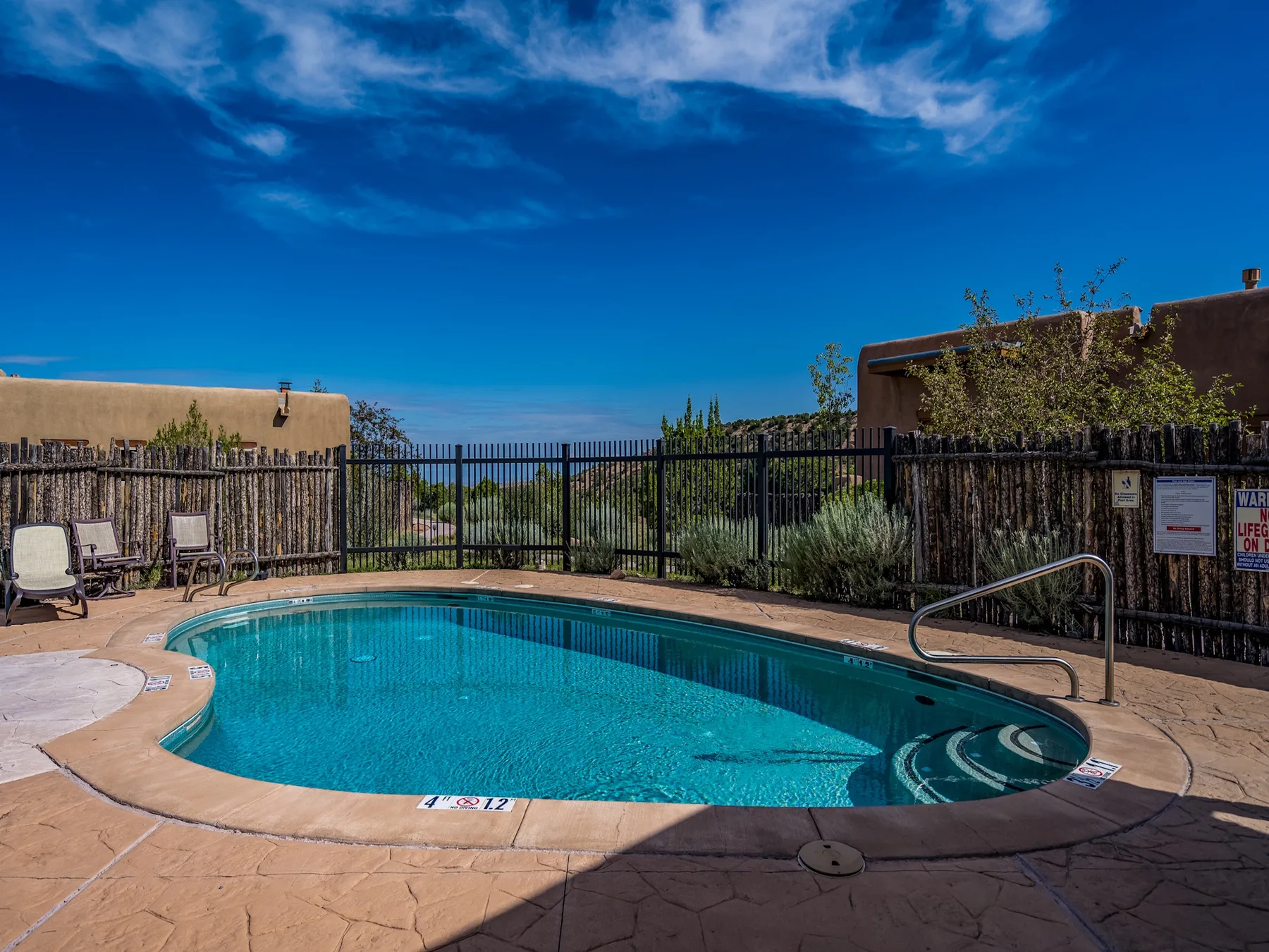An outdoor swimming pool sits beside a fenced patio under a bright blue sky.